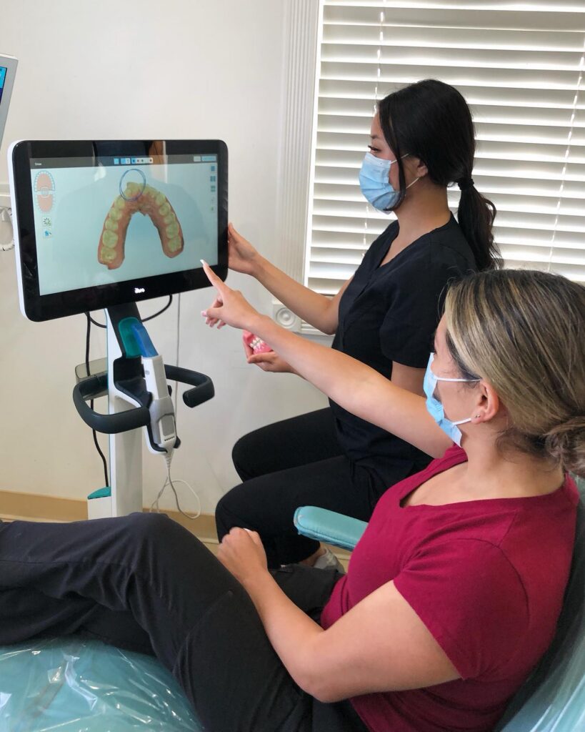 Dr. Christina Pak, wearing protective blue scrubs and a face mask, uses a dental model and handheld mirror to explain a procedure to a seated patient at Wellbeing Dentistry in Santa Clara, CA.