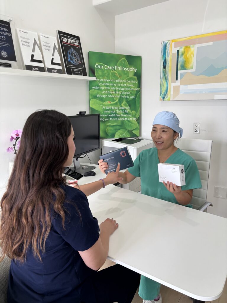 Dr. Christina Pak, wearing blue surgical scrubs, a face mask, and a head covering, explains a dental treatment to a seated patient using a physical mouth model at Well Being Dentistry in Santa Clara, CA. The patient holds a hand mirror and observes.