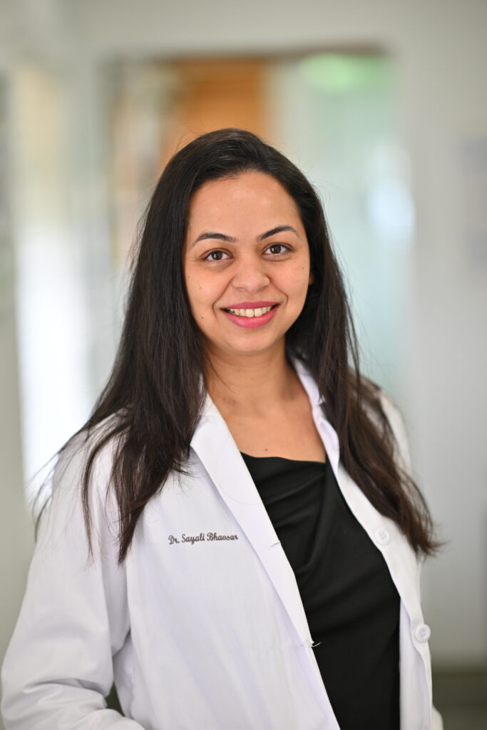 Professional headshot of Dr. Sayali Bhavsar, smiling warmly in a white lab coat at Wellbeing Dentistry.